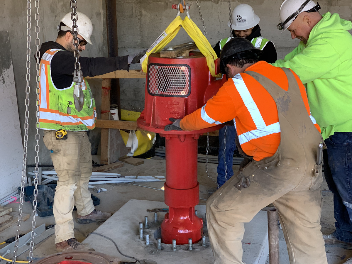 Crew guiding a red fire pump head onto its base with a chain hoist